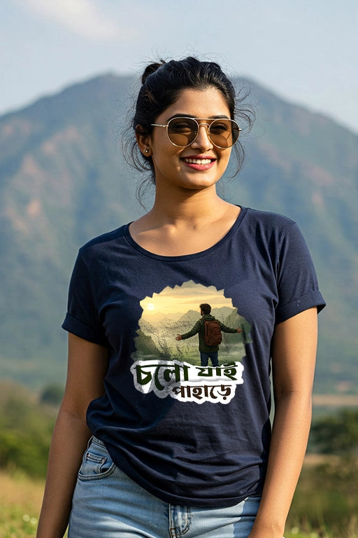 Woman wearing a navy blue t-shirt with a graphic design and text, standing in front of a mountainous landscape.